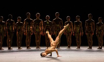 A dancer in a skin tight costume breakdances while a line of other dances watches from the shadows