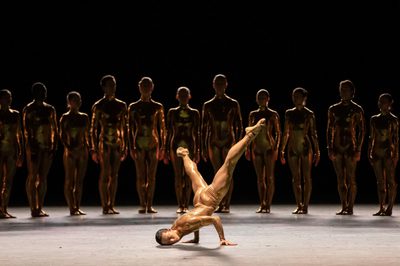 A dancer in a skin tight costume breakdances while a line of other dances watches from the shadows