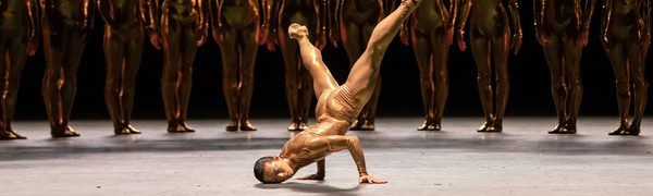 A dancer in a skin tight costume breakdances while a line of other dances watches from the shadows