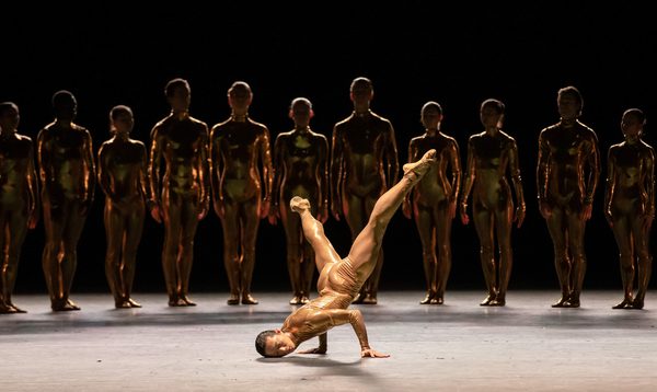 A dancer in a skin tight costume breakdances while a line of other dances watches from the shadows