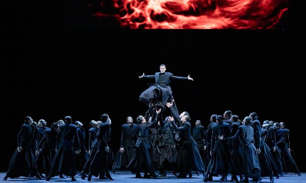 A man in a billowing black outfit is held up by a crowd of dancers in similar clothing. His arms are held out wide