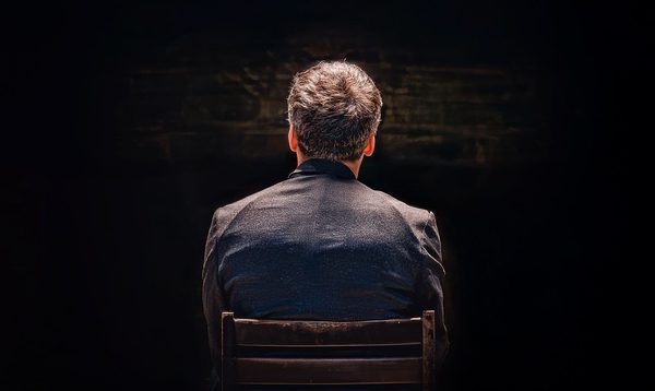 A man in a black suit is sat with his back to the viewer. He is lit by spotlight from above.