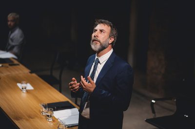 A middle aged man in a blue suit looks up with his hands held in front of him. There is a long wooden table beside him.