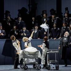 An early 20th century surgeon operates on someone in the middle of a crowded theatre. He holds up a piece of viscera in tongs to the shock of onlookers.