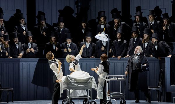 An early 20th century surgeon operates on someone in the middle of a crowded theatre. He holds up a piece of viscera in tongs to the shock of onlookers.
