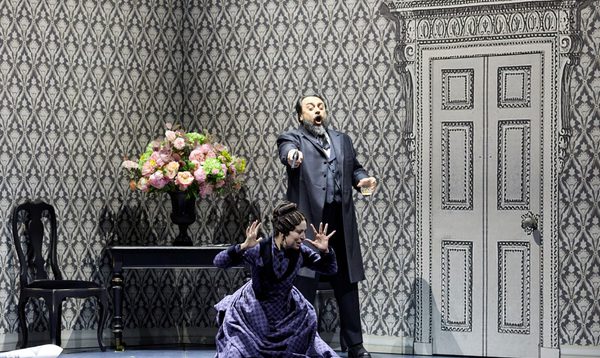 A man sings with a glass of alcohol in one hand and a gun in the other. He is pointing the gun at the head of a terrified woman kneeling in front of him. They are both dressed in early 20th century outfits