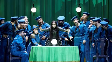 A fortune teller extends her hands forward as she sings. A crystal ball rests on the table in front of her. Around her are men dressed as early 20th century sailors.