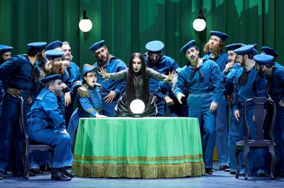 A fortune teller extends her hands forward as she sings. A crystal ball rests on the table in front of her. Around her are men dressed as early 20th century sailors.