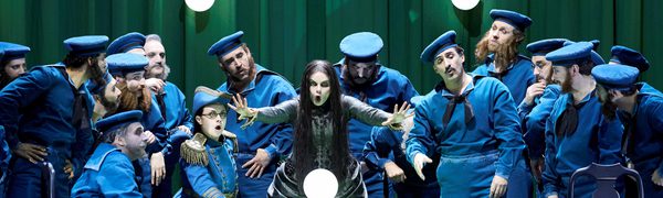 A fortune teller extends her hands forward as she sings. A crystal ball rests on the table in front of her. Around her are men dressed as early 20th century sailors.
