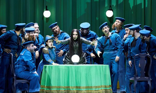 A fortune teller extends her hands forward as she sings. A crystal ball rests on the table in front of her. Around her are men dressed as early 20th century sailors.