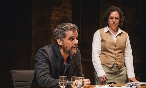 A middle-aged man in a black suit leans forward at a desk, speaking to someone out of frame. A woman in a shirt and vest stands next to him.