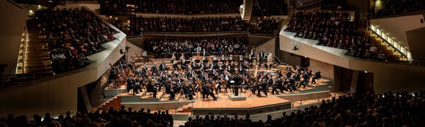 An orchestra performs in a modern concert hall. The audience is sat listening from all sides of the stage.