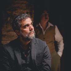 A middle-aged man in a black suit looks off to the side. A woman in a vest and shirt stands in the background behind him