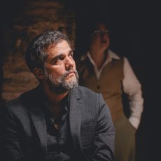 A middle-aged man in a black suit looks off to the side. A woman in a vest and shirt stands in the background behind him