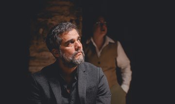 A middle-aged man in a black suit looks off to the side. A woman in a vest and shirt stands in the background behind him