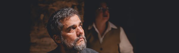 A middle-aged man in a black suit looks off to the side. A woman in a vest and shirt stands in the background behind him