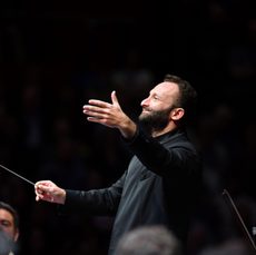 A bearded conductor smiles and holds his hand out