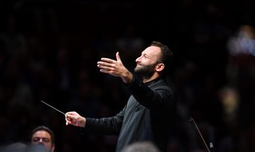 A bearded conductor smiles and holds his hand out