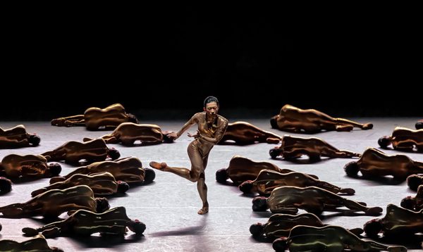 An androgynous dancer in a skin tight outfit poses with one leg raised. On the floor around them are many other people in similar outfits lying with their back to the viewer