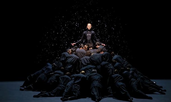 A woman in a shiny black outfit stands amidst a group of people in black who are crouched around her, their faces hidden. Snow falls around the central woman.