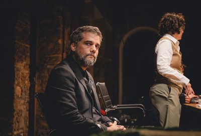 A middle aged man in black is sat looking at the viewer with a sad and weary expression. A woman in a brown vest is stood facing away next to him.