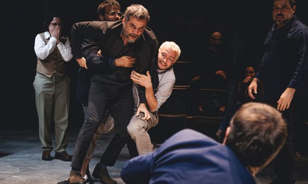 A middle aged man in a black suit is being held back by two other men. The man in black is lunging towards a man in a blue suit. Behind the scene a shocked woman covers her mouth.
