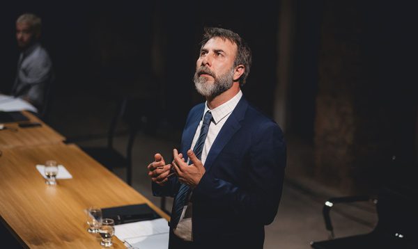 A middle aged man in a blue suit looks up with his hands held in front of him. There is a long wooden table beside him.