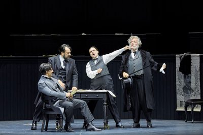 Four people in grey, early 20th century formal outfits are gathered around a table. A woman in a vest sings while extending her fist towards an older man with a cane.