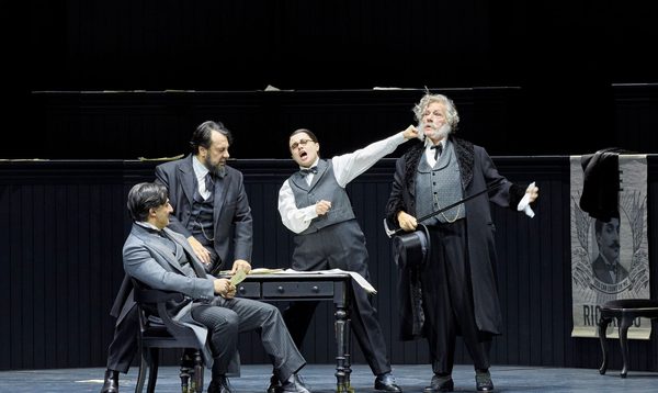 Four people in grey, early 20th century formal outfits are gathered around a table. A woman in a vest sings while extending her fist towards an older man with a cane.