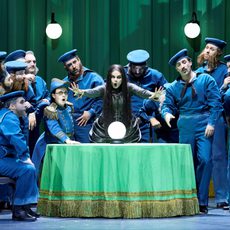 A fortune teller extends her hands forward as she sings. A crystal ball rests on the table in front of her. Around her are men dressed as early 20th century sailors.