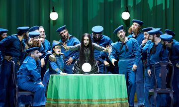 A fortune teller extends her hands forward as she sings. A crystal ball rests on the table in front of her. Around her are men dressed as early 20th century sailors.