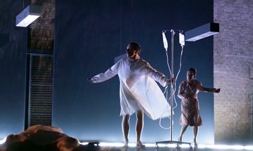 A person lies on the floor while another person stands in a hospital gown holding an IV pole. A distressed woman stands behind them.