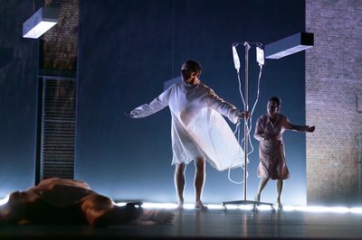 A person lies on the floor while another person stands in a hospital gown holding an IV pole. A distressed woman stands behind them.