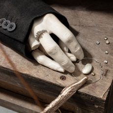 A clay sculpture hand sits on a wooden box with some fingers broken off and wearing a ring surrounded by small trinkets