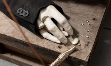 A clay sculpture hand sits on a wooden box with some fingers broken off and wearing a ring surrounded by small trinkets