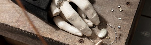 A clay sculpture hand sits on a wooden box with some fingers broken off and wearing a ring surrounded by small trinkets