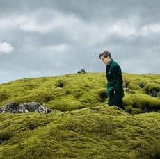 A man in a green jacket walking across a mossy hill