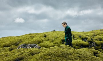 A man in a green jacket walking across a mossy hill