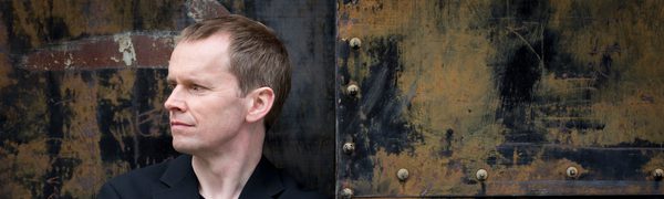 A man dressed in black leaning against a distressed looking door while looking off to the side