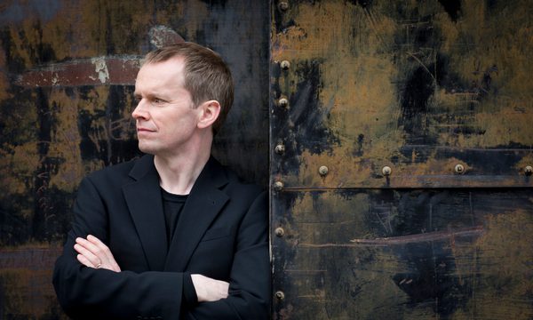 A man dressed in black leaning against a distressed looking door while looking off to the side