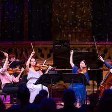 A group of string musicians on stage, holding their bows in the air