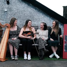 four women sit on a bench outside with their instruments, clarsach, fiddle, whistle and keyboard