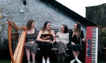 four women sit on a bench outside with their instruments, clarsach, fiddle, whistle and keyboard