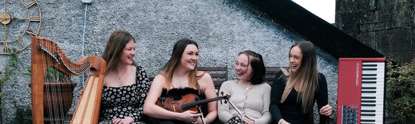 four women sit on a bench outside with their instruments, clarsach, fiddle, whistle and keyboard