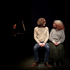 Two people sit illuminated on a dark stage while turning to look at each other