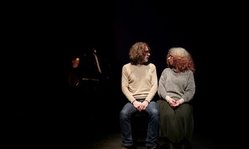 Two people sit illuminated on a dark stage while turning to look at each other
