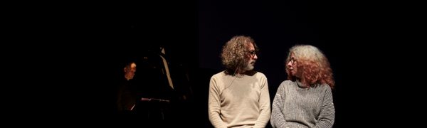 Two people sit illuminated on a dark stage while turning to look at each other
