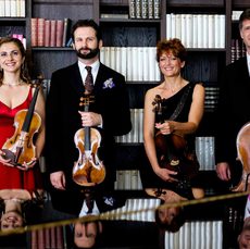 A quartet of musicians standing in front of a bookcase holding their instruments