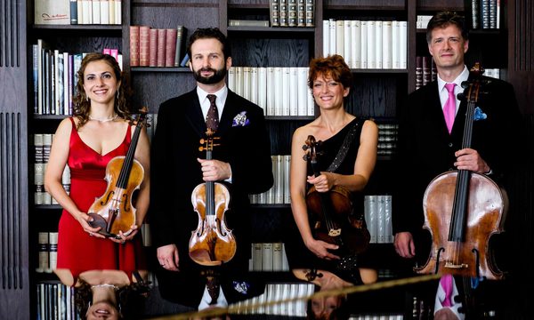 A quartet of musicians standing in front of a bookcase holding their instruments