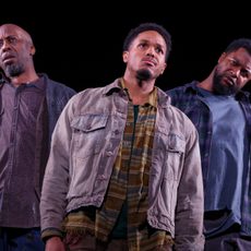 Three men stand on stage with serious expressions all looking in different directions